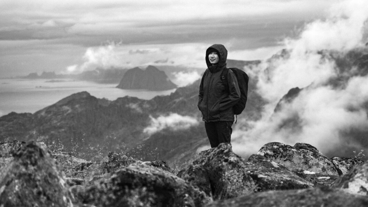 ノルウェー・ノールランド地方の山岳地帯で、フード付きの技術的なアウトドアジャケットとバックパックを身につけて岩場に立つ人物(Norrønaが拠点とするノルウェーの山岳環境のイメージ)