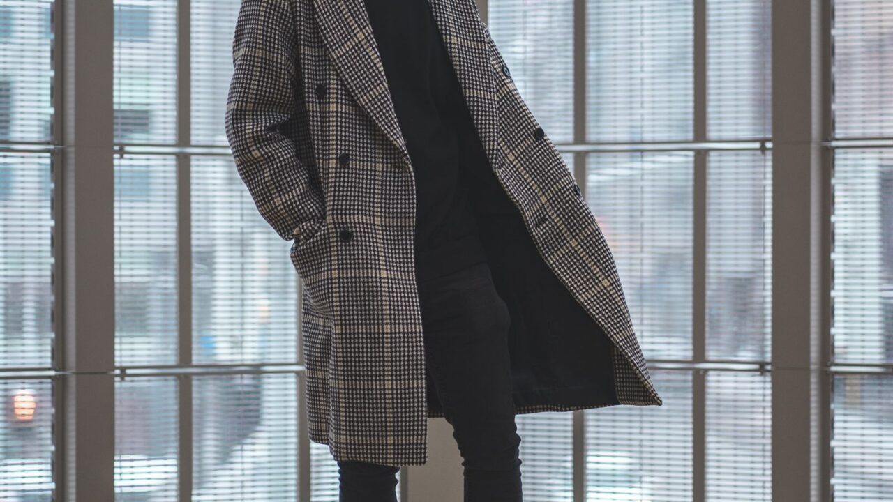 houndstooth柄のオーバーサイズコートを着た男性が窓辺に佇む写真(Hed Maynerの誇張されたプロポーションのイメージ)