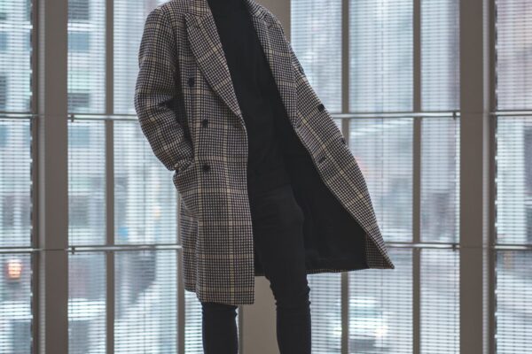 houndstooth柄のオーバーサイズコートを着た男性が窓辺に佇む写真(Hed Maynerの誇張されたプロポーションのイメージ)