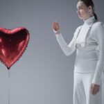 白いミニマルなニットとハーネスを着た女性が赤いハート型バルーンを見つめている。AMI Parisの赤いハートロゴ「Ami de Coeur」と気負わない上質さという世界観のイメージ