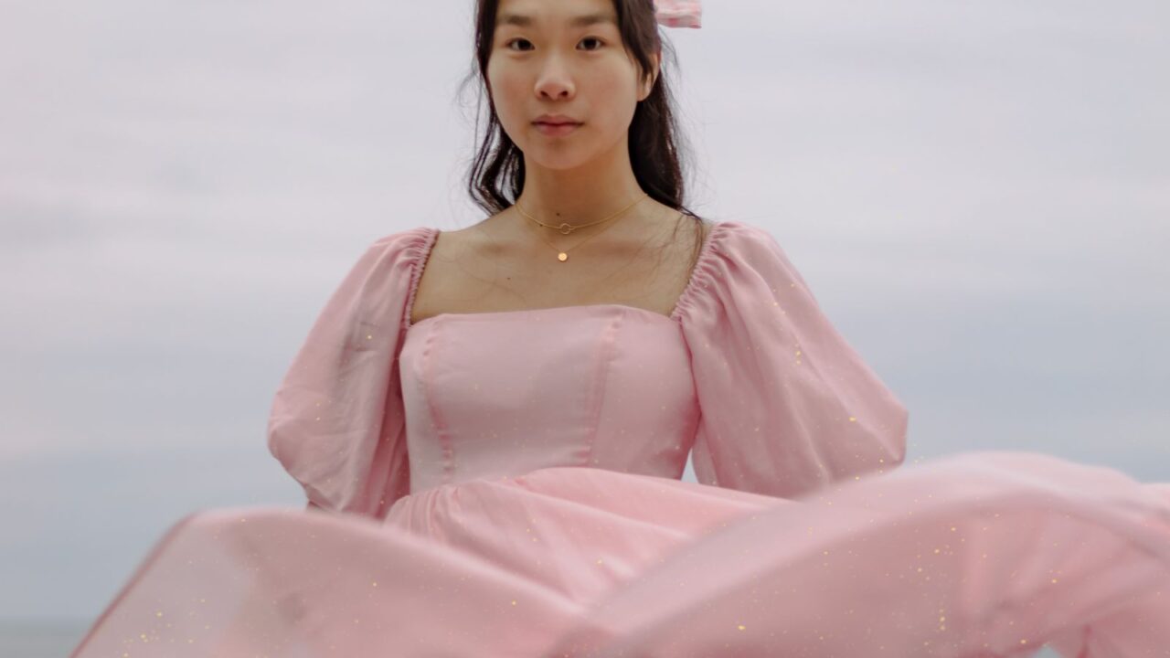 海辺を背景に、ボリュームのあるパフスリーブとふくらんだシルエットのピンクのドレスを着た人物(Cecilie Bahnsenのバブルドレスの意匠イメージ)