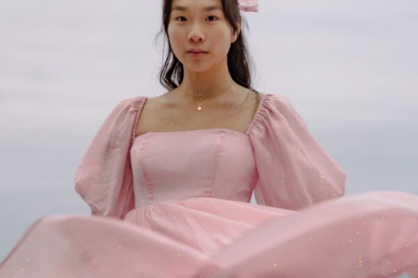 海辺を背景に、ボリュームのあるパフスリーブとふくらんだシルエットのピンクのドレスを着た人物(Cecilie Bahnsenのバブルドレスの意匠イメージ)