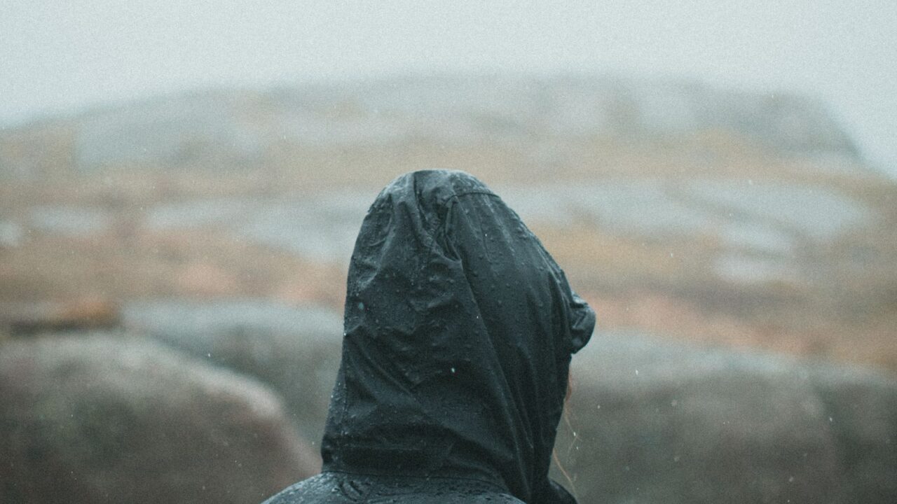 黒い防水シェルジャケットを着た人物の後ろ姿、雨に濡れた岩山(Arc'teryxのテクニカルアウター世界観イメージ)