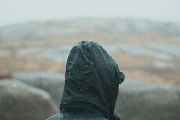 黒い防水シェルジャケットを着た人物の後ろ姿、雨に濡れた岩山(Arc'teryxのテクニカルアウター世界観イメージ)